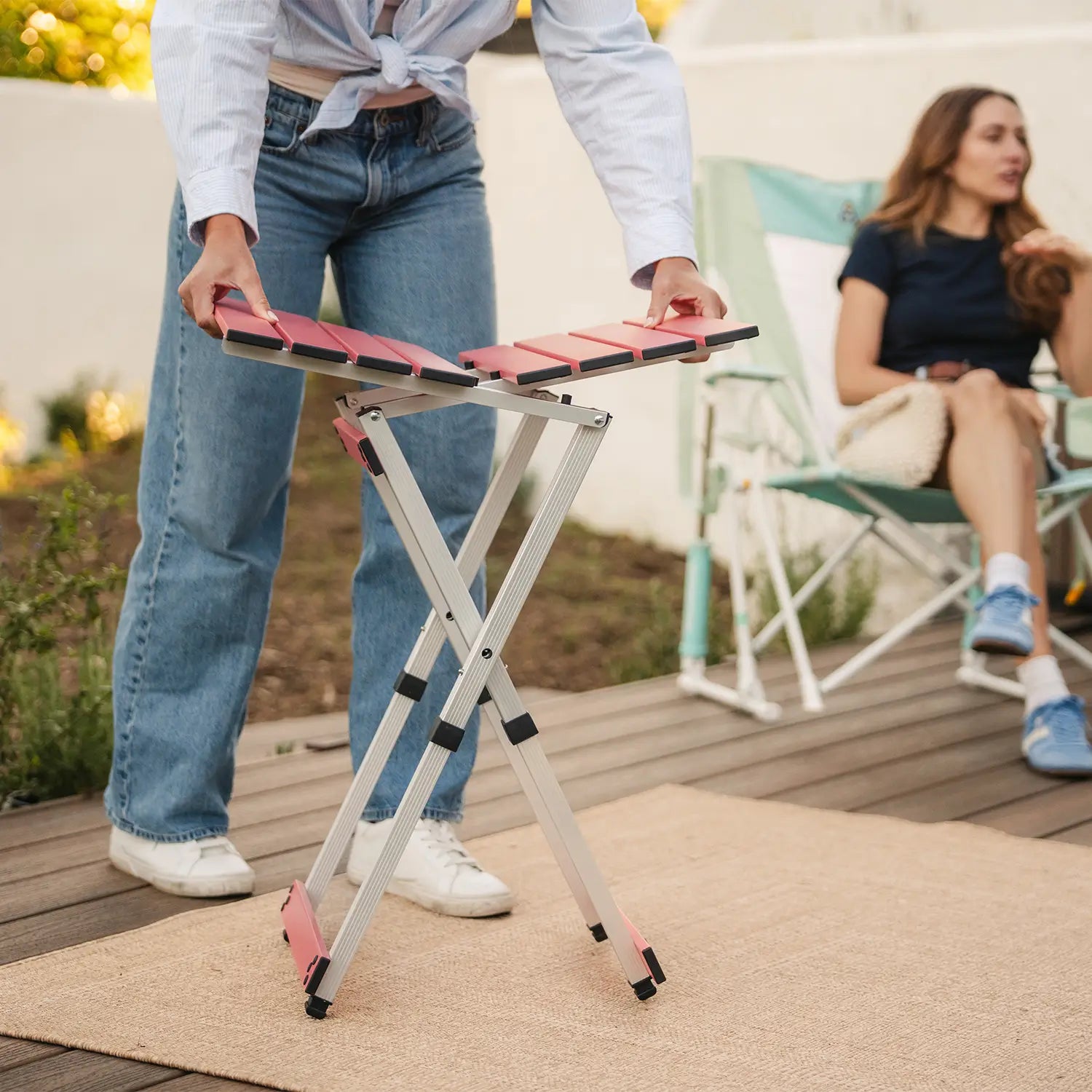 A woman assembling a coral Compact Camp Table near a group sitting in rockers. 