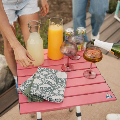 A woman pouring a glass of juice into a cup thats positioned on a coral Compact Camp Table. 