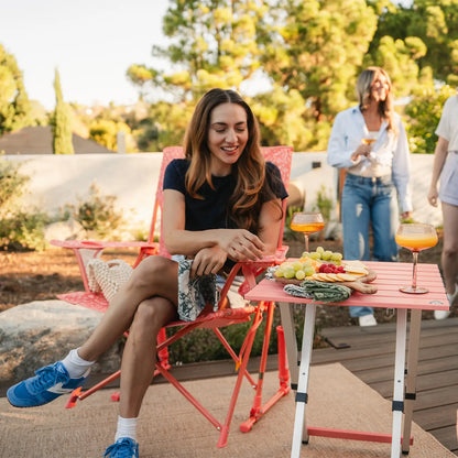 A woman sitting in a coral Comfort Pro Rockers is grabbing a snack resting on a coral Compact Camp Table. 