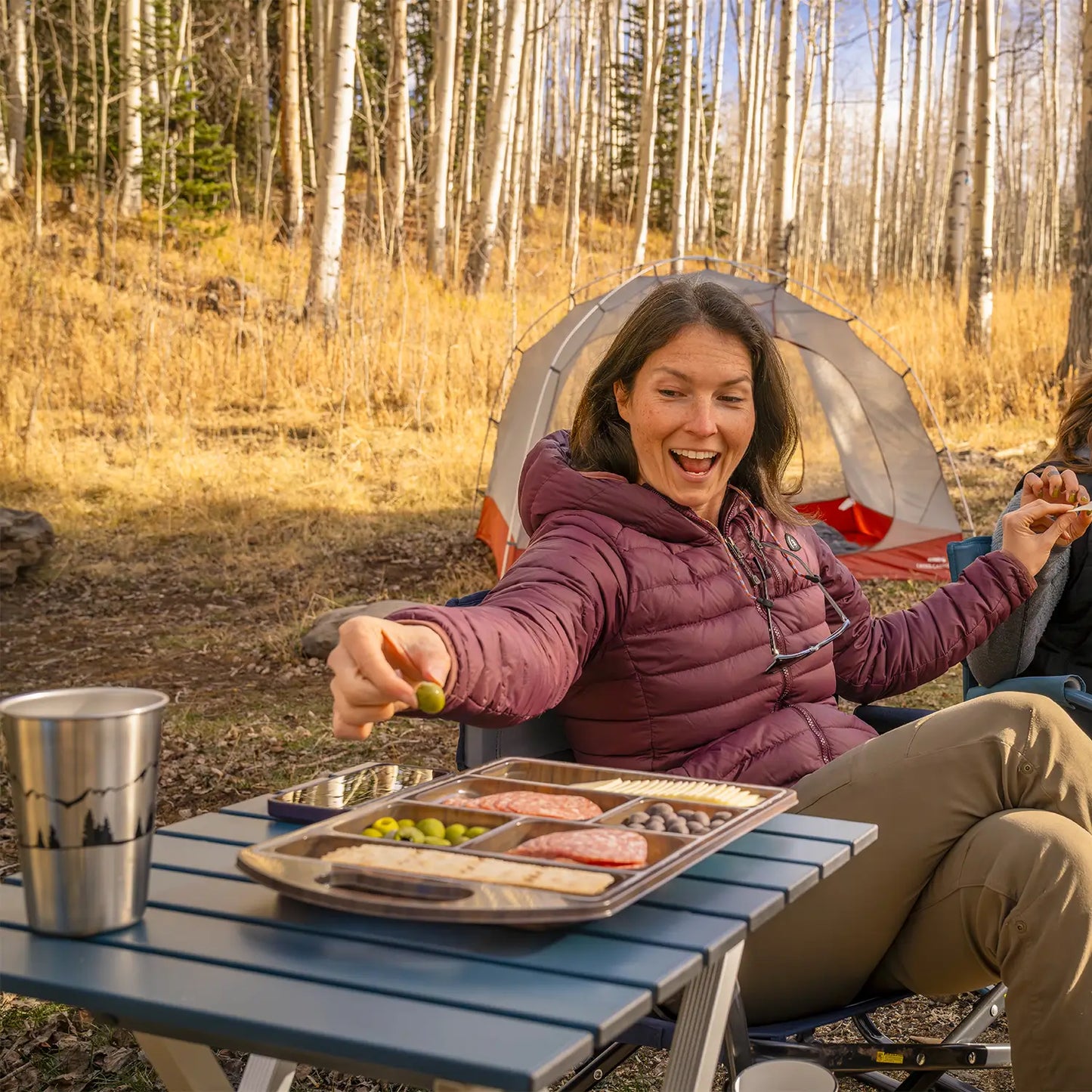 A woman reaching for an olive from a charcuterie tray thats resting on a teal Compact Camp Table. 
