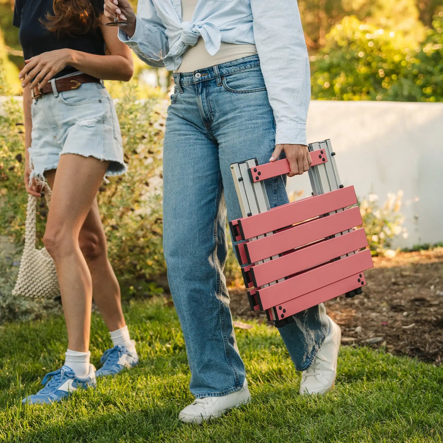 Two women walking in a backyard setting while one woman carries a coral Compact Camp Table. 