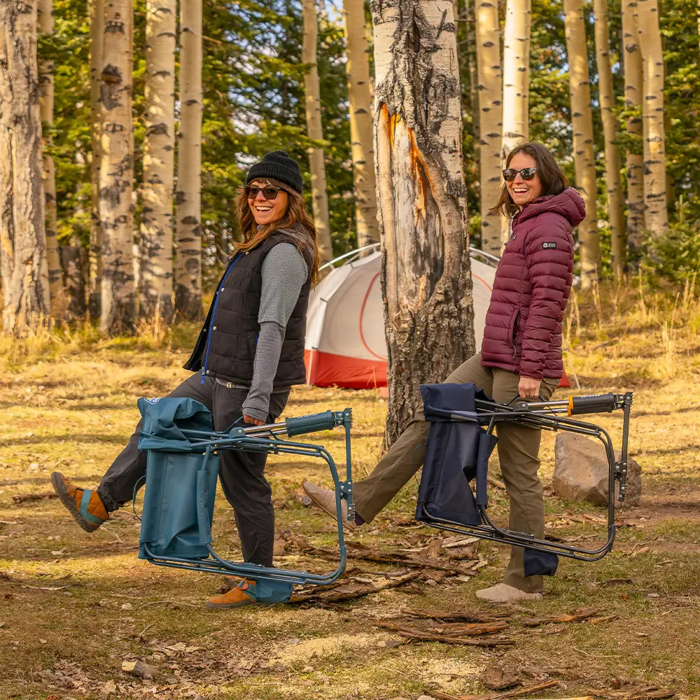 Two people carrying folded Teal Grab & Go Rockers through a wooded campsite.